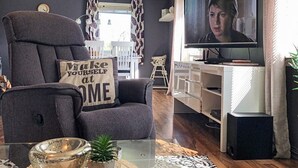 Een moderne woonkamer met een grijze fauteuil, een glazen salontafel, een televisie en een boekenkast.