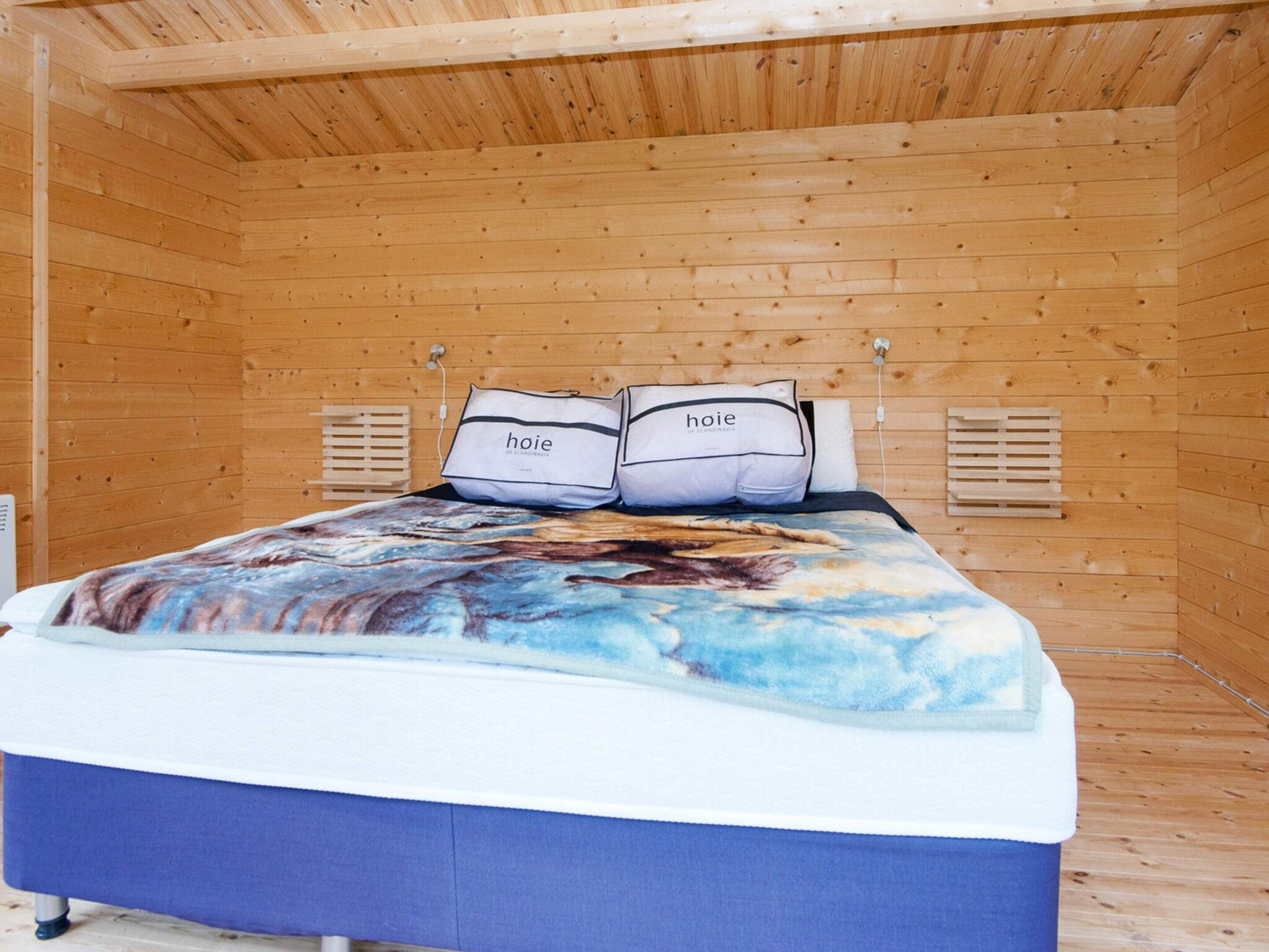Ein Zimmer mit Holzboden, einem Bett, zwei Kissen mit dem Schriftzug 'hoie' und einem Fenster mit Jalousien.