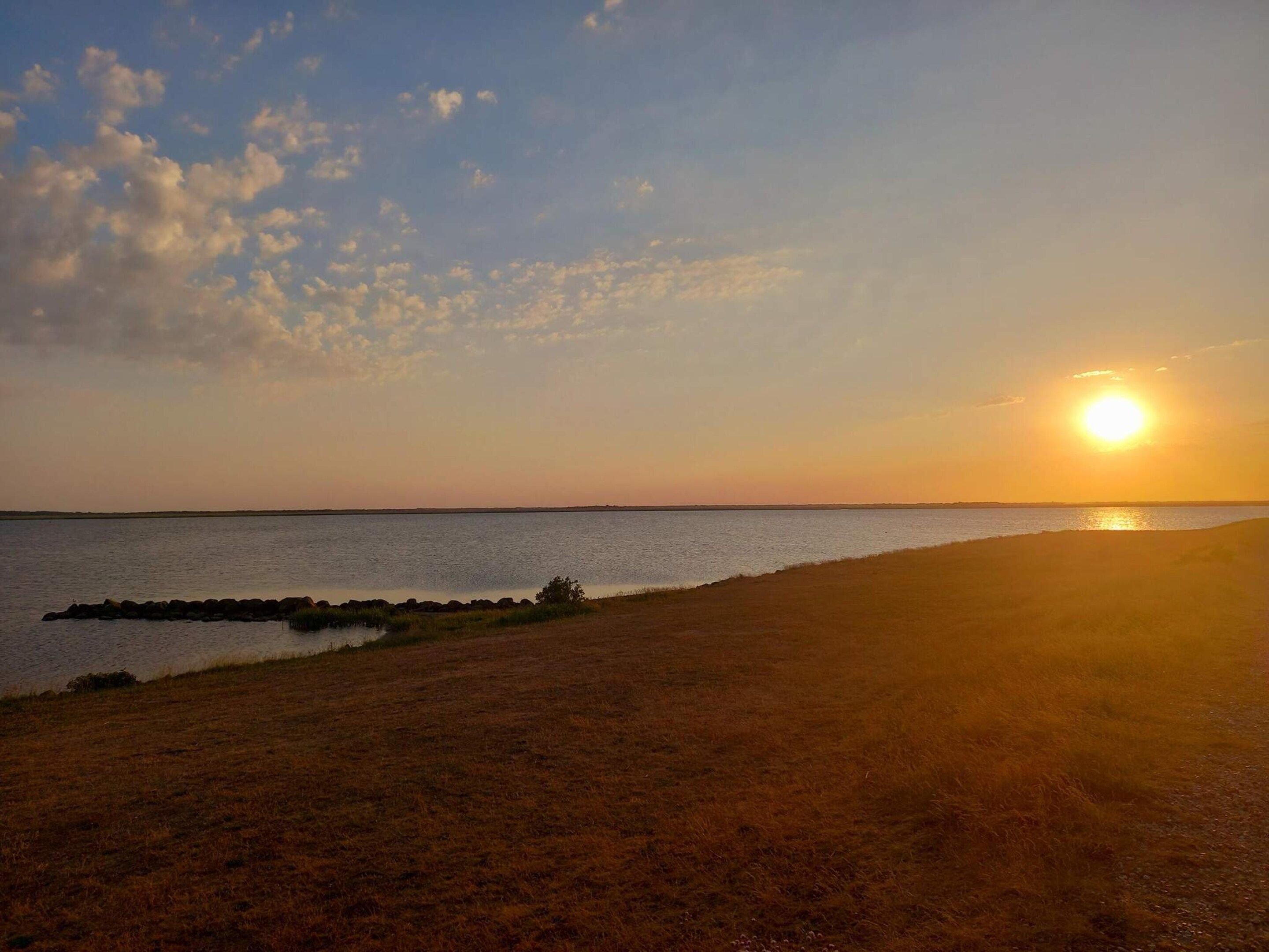 Auringonlasku vesistön yllä, edessä hiekkaranta ja kallioinen niemeke.