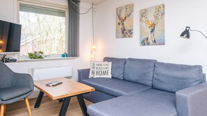 A modern living room with a grey sofa, a wooden coffee table, a grey armchair, and a television. There are two paintings on the wall and a lamp on the right.