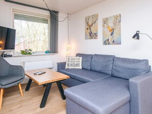 A modern living room with a grey sofa, a wooden coffee table, a grey armchair, and a television. There are two paintings on the wall and a lamp on the right.