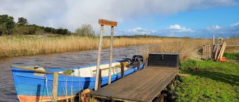 A wooden dock with a blue boat moored at the end, a small shed, and a grassy bank with reeds.