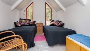 A small, cozy room with two beds, a wooden dresser, a wicker chair, and a large window.