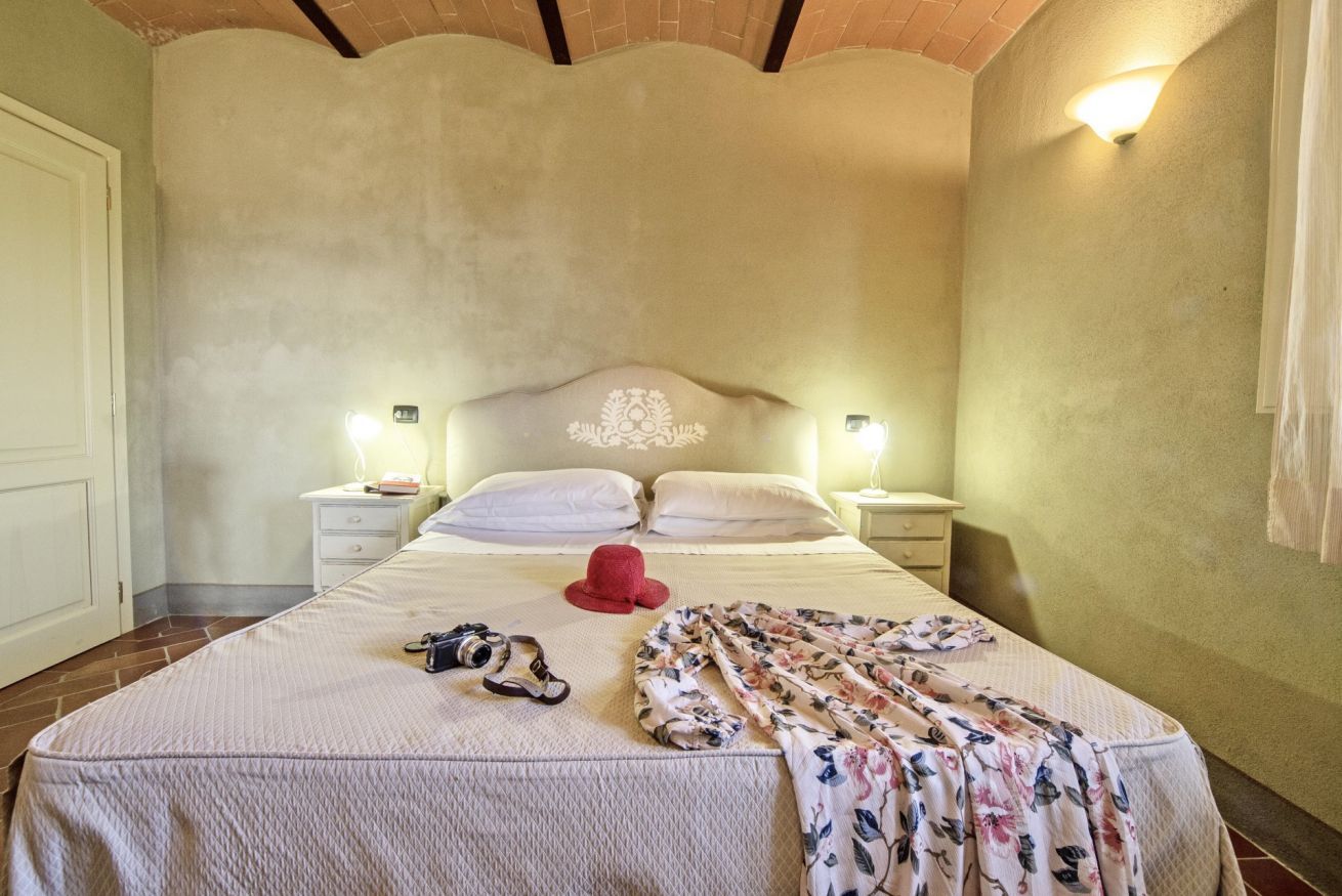 A bedroom with a bed, two bedside tables, a nightstand, a red hat, a floral blanket, and a pair of sandals.