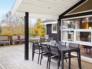A covered outdoor seating area with tables and chairs, a wooden deck, and a view of a building with large windows. - 10 Person Holiday Home in Hojby (Hojby)