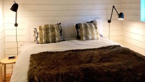 A bedroom with a large bed, two pillows, a blanket, a bedside table with a lamp, and a wall-mounted lamp. - 4 Person Holiday Home in Garphyttan (Garphyttan)