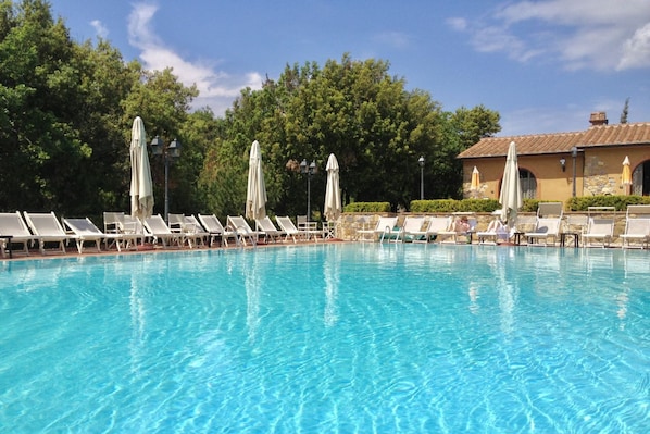 A swimming pool with lounge chairs and umbrellas, surrounded by trees and a building with a tiled roof.