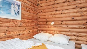 Une chambre confortable, aménagée dans le style d’un chalet, avec des murs en bois, un lit recouvert d’une literie blanche, un plaid de couleur moutarde et une table de chevet avec une lampe.