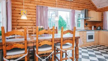 A wooden cabin interior with a dining table, chairs, a kitchen area, and a view of the outdoors.