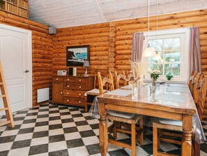 A log cabin interior with a checkered floor, wooden walls, a dining table with chairs, a window with curtains, and a painting on the wall. - 6 Person Holiday Home in Hovborg (Hovborg)