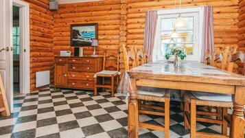 A log cabin interior with a checkered floor, wooden furniture, a dining table, chairs, a dresser, a painting, a lamp, and a window with curtains.