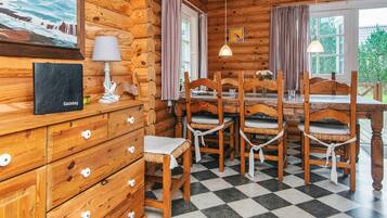 A log cabin interior with a checkered floor, wooden furniture, a dining table with chairs, a lamp, a framed picture, and a window with curtains.