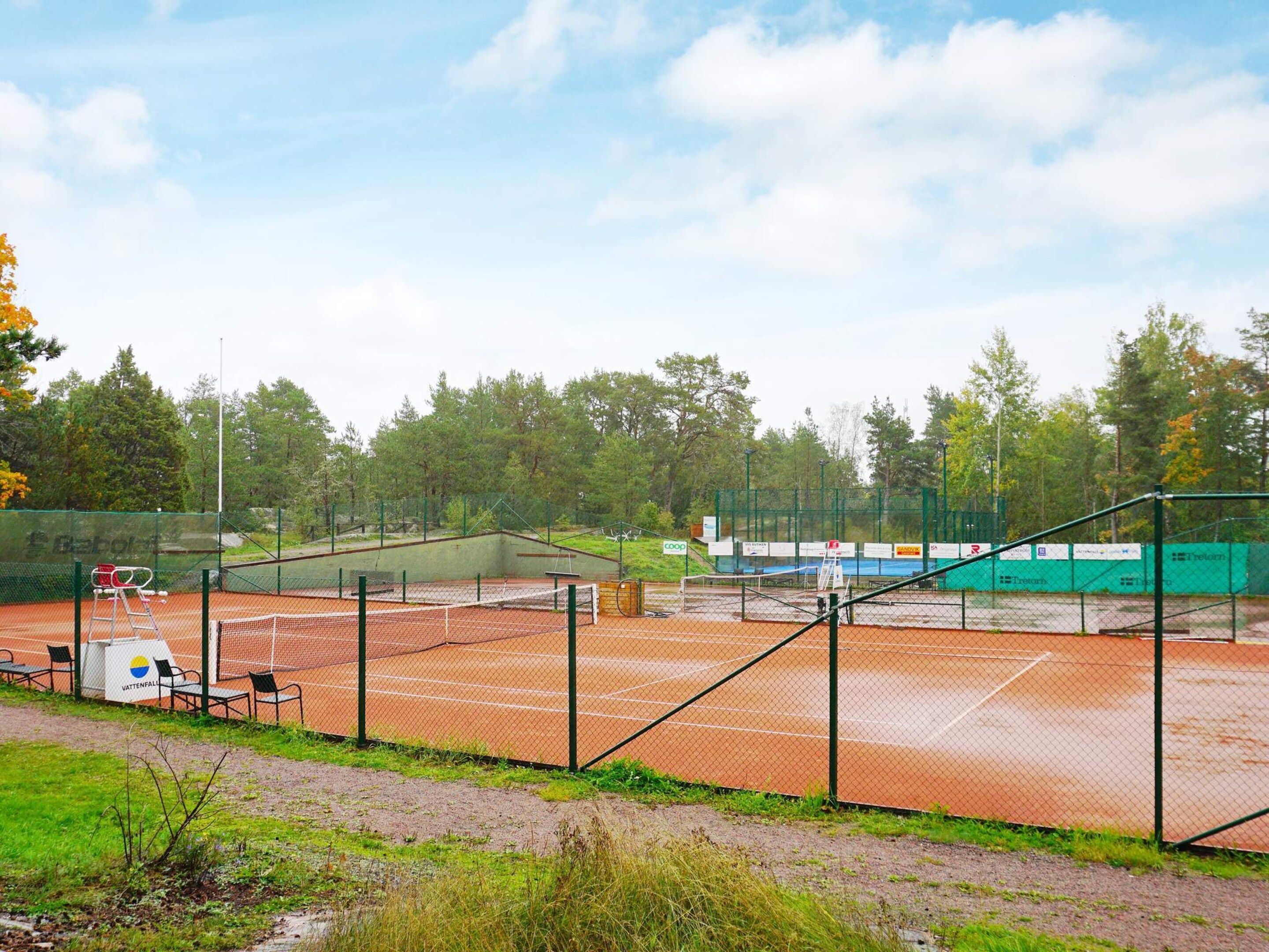 An outdoor tennis court with a net, surrounded by a green fence, and trees in the background.