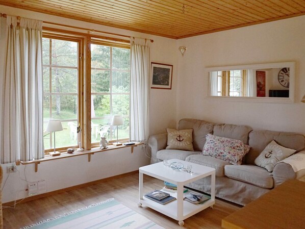 Una sala de estar acogedora con techo de madera, un sofá blanco con cojines estampados, una mesa de centro blanca con libros y ventanas amplias con cortinas.
