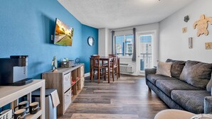 A modern living room with a blue accent wall, a dining area with wooden furniture, and a grey sofa. - NEW! 10th Floor! Tip-Sea Turtle, Beach View! (Myrtle Beach)