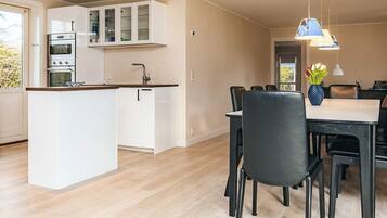 A modern kitchen with white cabinetry, a built-in oven, and a central island. There is a dining area with a table and chairs, and a vase of flowers on the table.