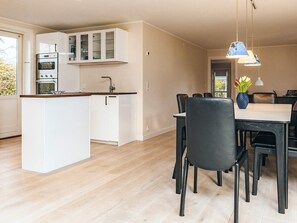 Une cuisine moderne avec des meubles blancs, un four encastré et un îlot central. On y trouve un coin repas avec une table et des chaises, ainsi qu'un vase de fleurs posé sur la table.