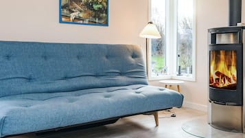 A modern living room with a blue sofa, a painting on the wall, a window with a view, and a wood-burning stove.