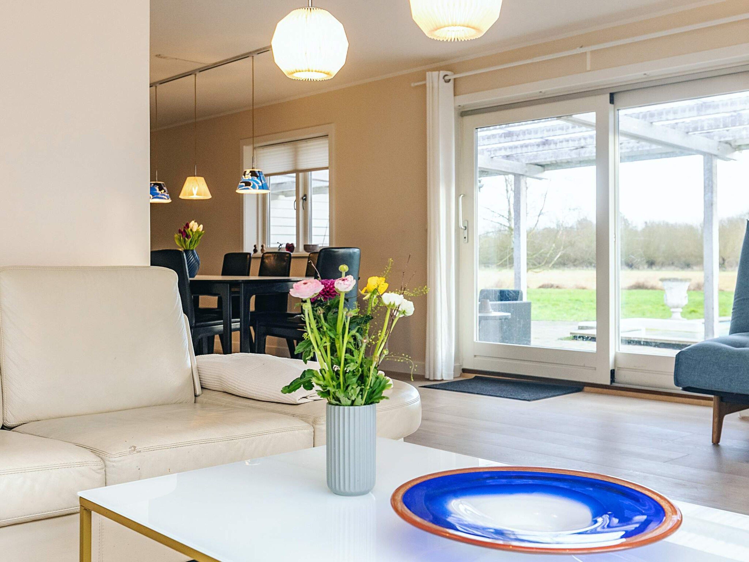 A living room with a sofa, a coffee table with a vase of flowers, and a dining area in the background.