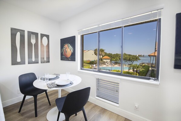 A dining area with a round table, two chairs, and a view of a pool and buildings through a large window.