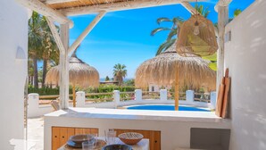 A poolside area with a table set for dining, a thatched-roof structure, and a clear view of palm trees and a blue sky. - CostaBlancaDreams - Casa Juleo in Benissa (Benissa)