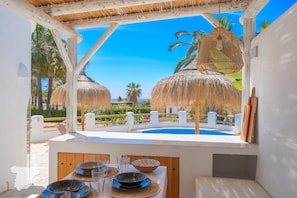 A poolside area with a table set for dining, a thatched-roof structure, and a clear view of palm trees and a blue sky. - CostaBlancaDreams - Casa Juleo in Benissa (Benissa)