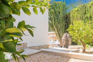 Un jardín con diversas plantas, incluyendo cactus y un árbol, un muro de piedra y una zona de piscina.