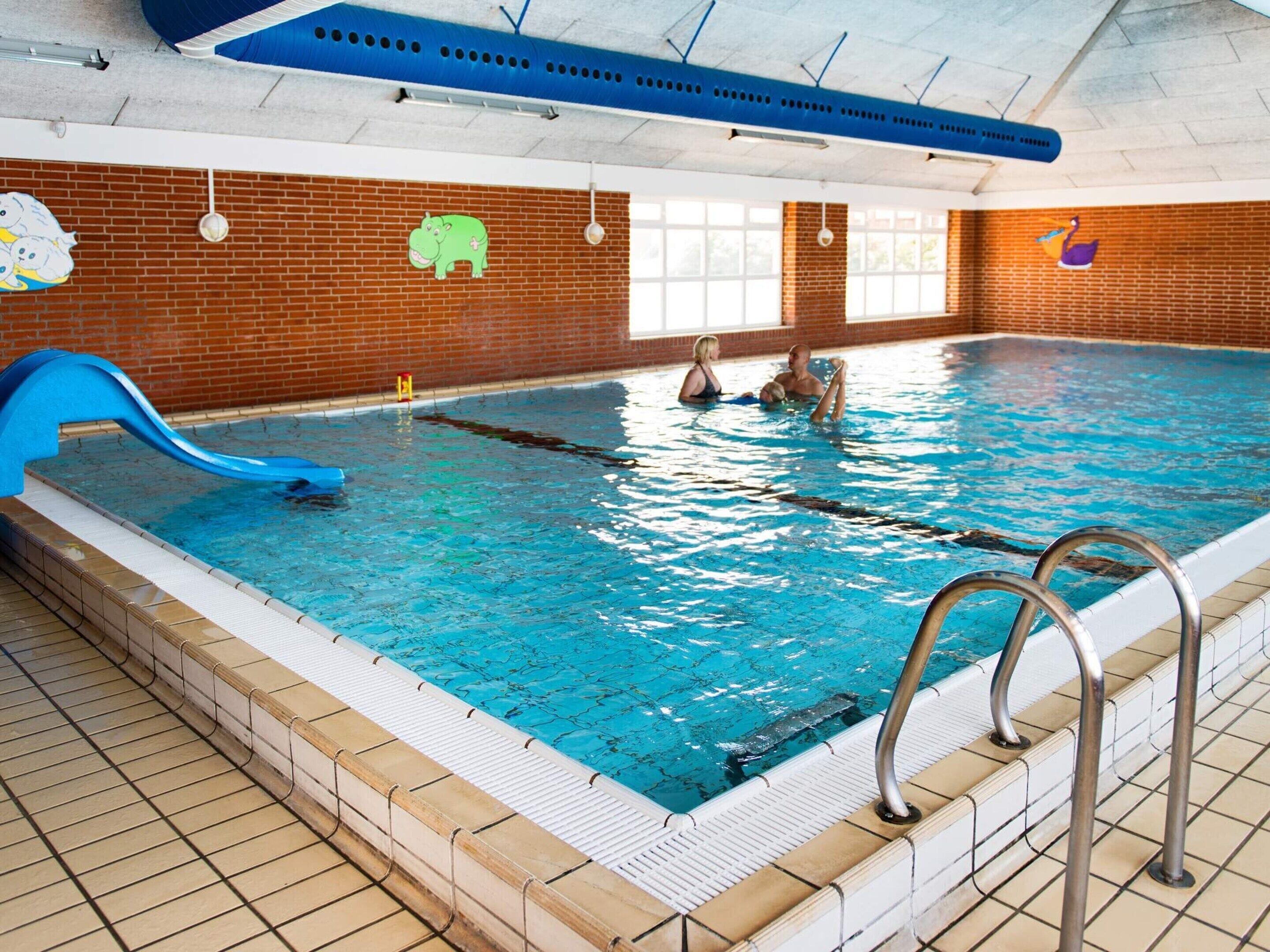 Piscina interna com um escorregador azul, parede de tijolos com murais de animais e chão de azulejos.