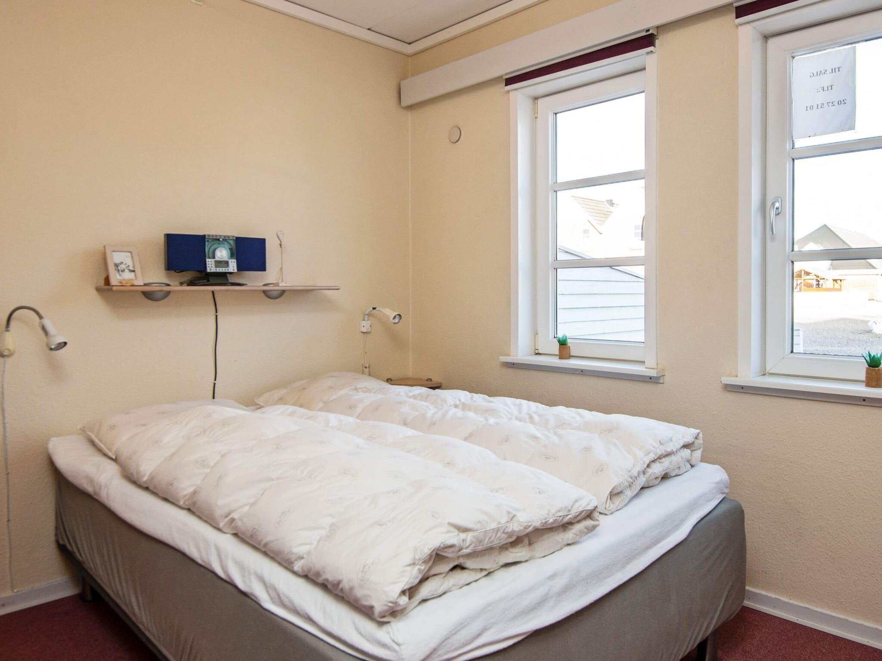 Una cama individual con un edredón blanco en una habitación con paredes color crema, una ventana con vistas a casas y una estantería con un marco de foto y una planta pequeña.