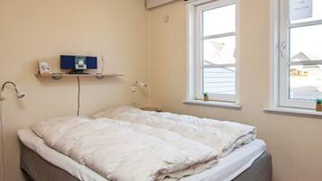 Una cama individual con un edredón blanco en una habitación con paredes color crema, una ventana con vistas a casas y una estantería con un marco de foto y una planta pequeña.