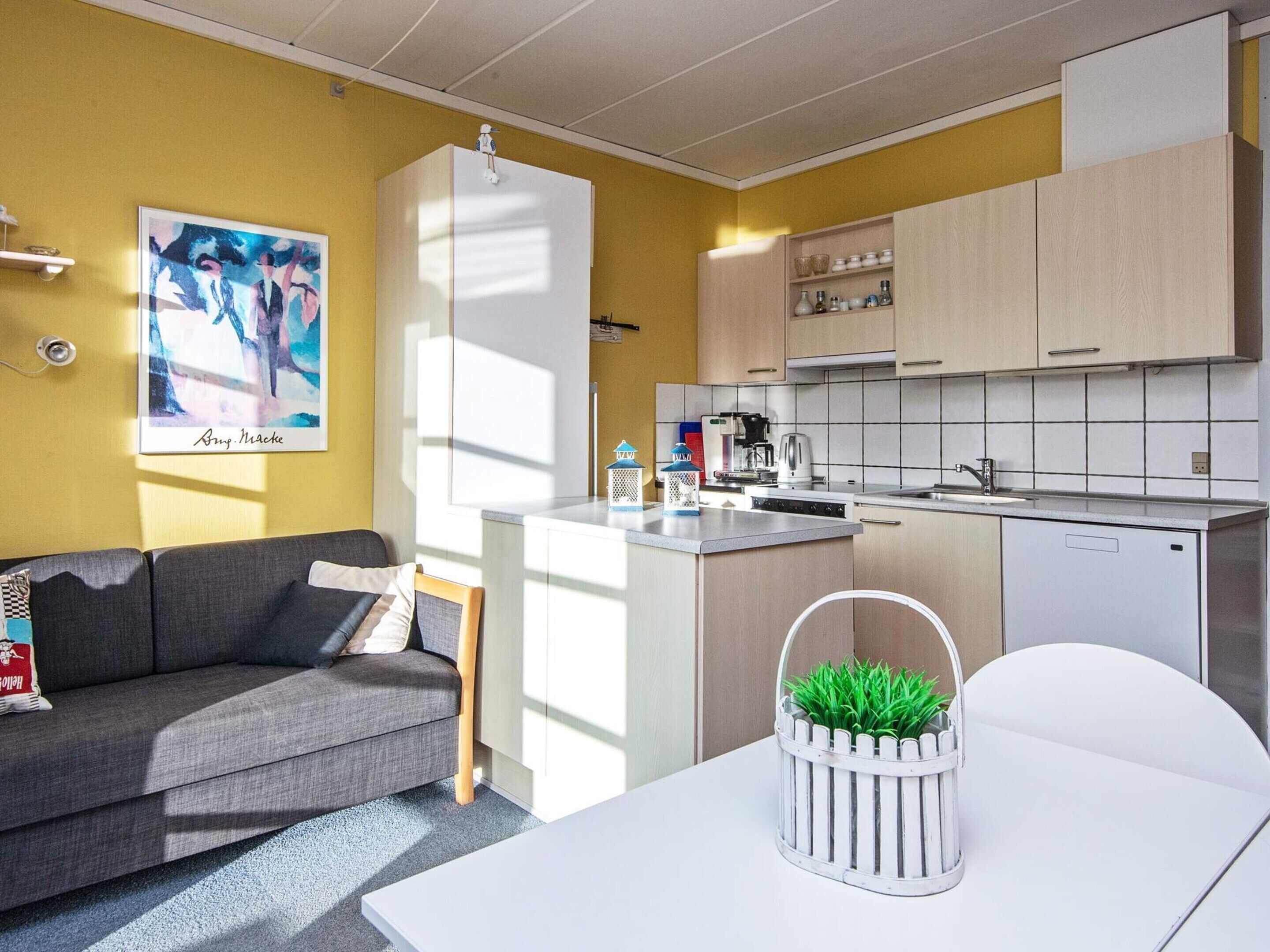 Ein kompakter Wohnraum mit Küchenzeile, einem grauen Sofa, einem kleinen Esstisch und einem weißen Schrank.