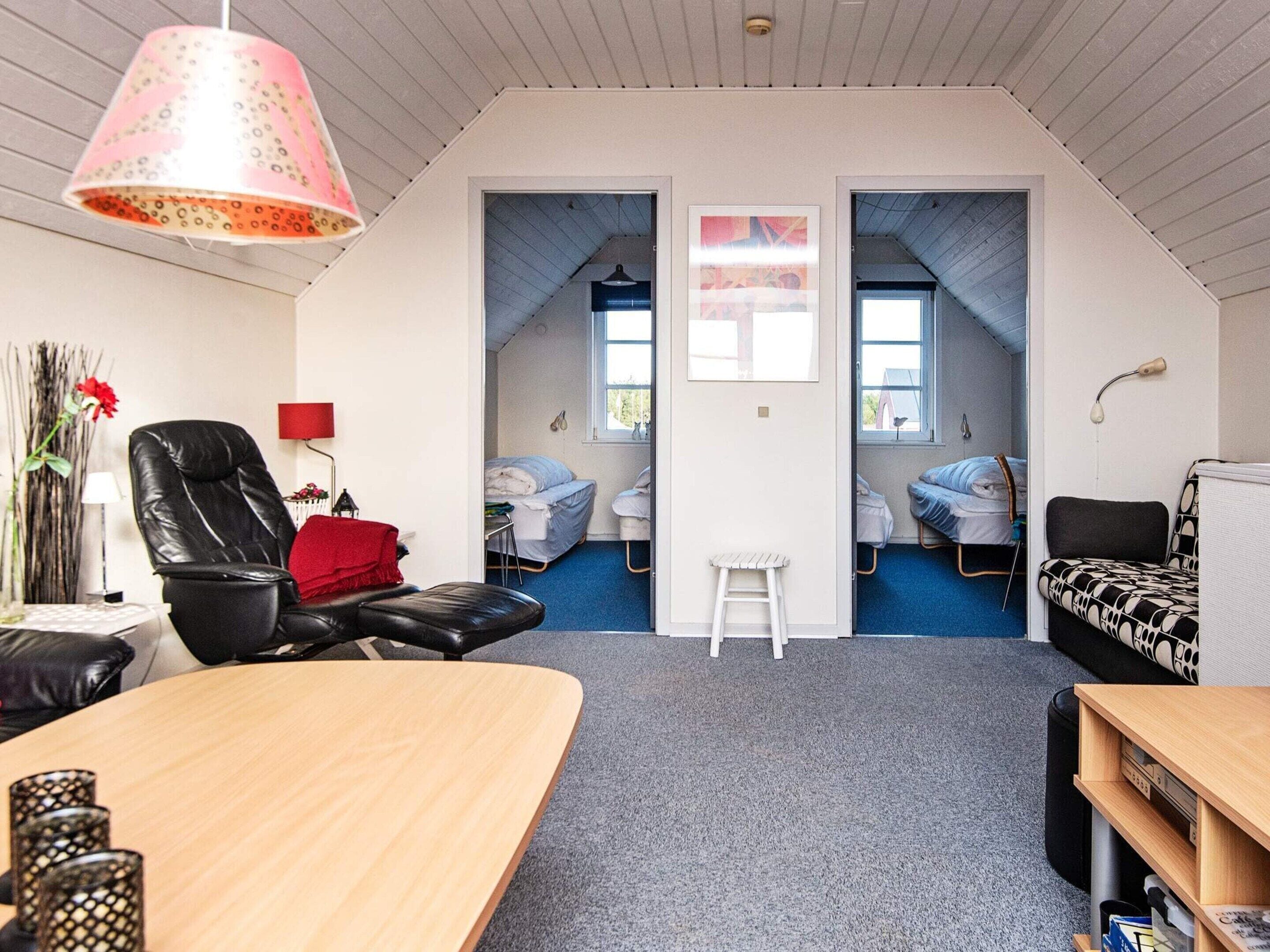 A room with a wooden desk, a black leather chair, a red pillow, a patterned sofa, and a small white stool. There are two beds with blue bedding and a ladder.