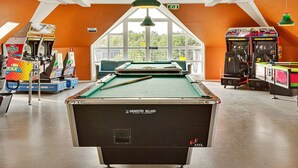 Mesa de bilhar numa sala com paredes laranja, jogos de fliperama e vista através de grandes janelas.
