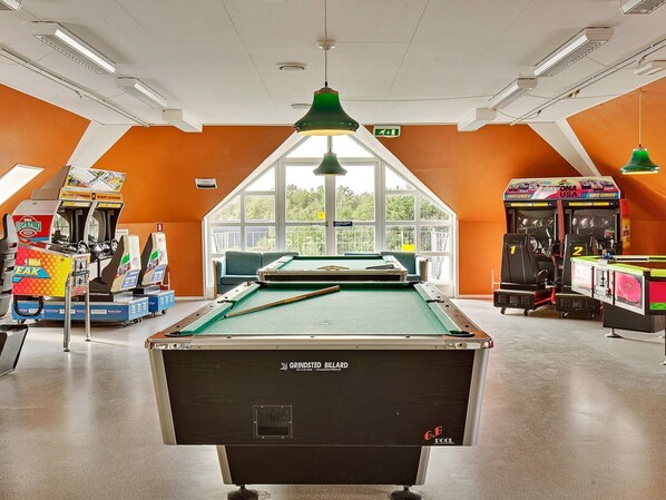 A pool table in a room with orange walls, arcade games, and a view through large windows.