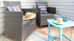 Deux chaises en osier ornées de coussins à rayures, une petite table avec un muffin et une tasse, et un chapeau de paille posé sur le sol.