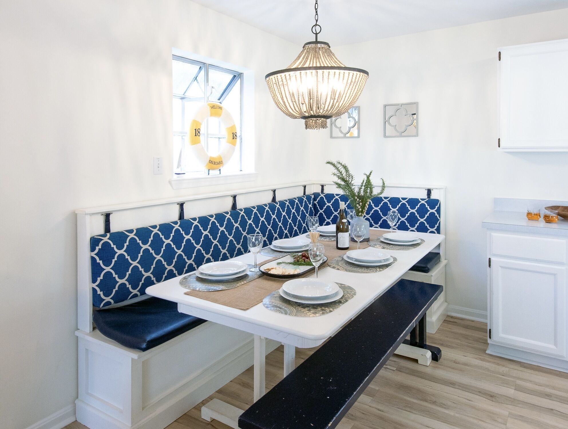 A dining area with a bench, a table set with plates and glasses, a chandelier, and a window with a 'WELCOME' sign.