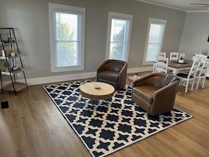 Een woonkamer met een kamerkleed met patroon, twee bruine leren fauteuils, een ronde houten salontafel, een boekenkast en een eethoek met witte stoelen en een tafel.