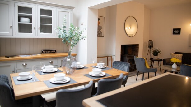 A dining area with a wooden table set for four, a vase with a plant, and a kitchen with white cabinets and a sign that reads "NO RIFF RAFF". - York Minster-3 bed-Sleeps 6-Fireplace-Free Parking (York)