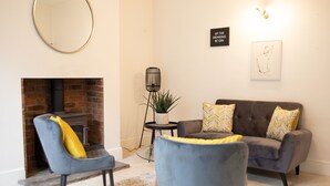 A living room with a fireplace, two armchairs, a sofa with patterned cushions, a round mirror, a wall-mounted light fixture, and a small table with a plant. - York Minster-3 bed-Sleeps 6-Fireplace-Free Parking (York)