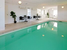Una piscina con superficie de agua cristalina, rodeada de paredes blancas y un techo con iluminación empotrada. Hay sillas y mesas negras dispuestas alrededor de la zona de la piscina.