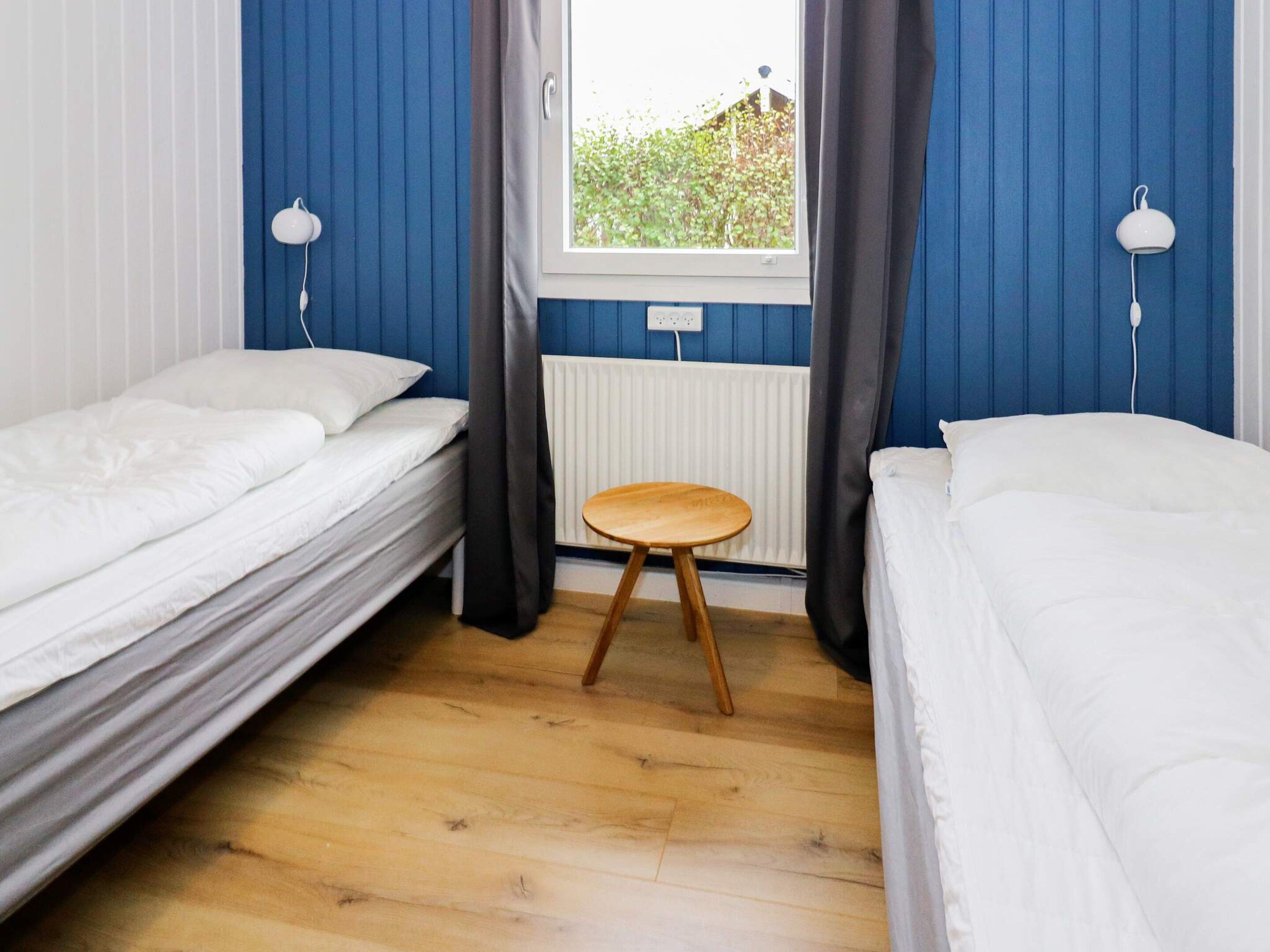 A room with two single beds, a wooden stool, a window with curtains, and a radiator.
