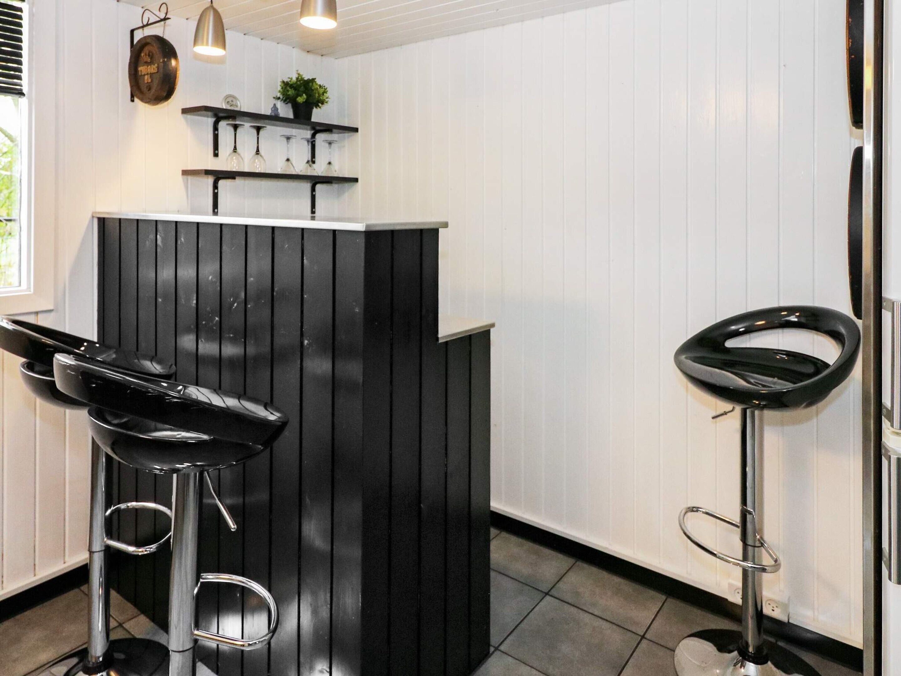 A bar area with two black bar stools, a shelf with wine glasses, and a hanging light fixture.