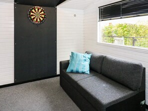 A room with a dartboard, a grey sofa with a blue pillow, and a window with a view of greenery.