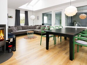 A modern living room with a fireplace, a sofa, a dining table with chairs, and a large window. - 4 Star Holiday Home in Hadsund (Hadsund)