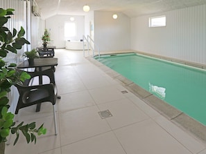 An indoor swimming pool with a tiled floor, a seating area with chairs and a table, and a window with blinds.