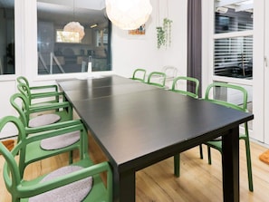 A dining area with a long table and green chairs, a window with blinds, and a hanging plant.