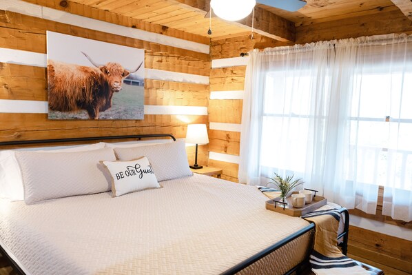 Una habitación de madera con una cama, una lámpara, un ventanal con cortinas y un cuadro de un toro.