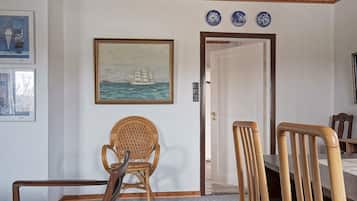 Una habitación con una mesa de comedor, dos sillas, una silla de mimbre, un cuadro de un barco en la pared y tres platos azules y blancos sobre la puerta.
