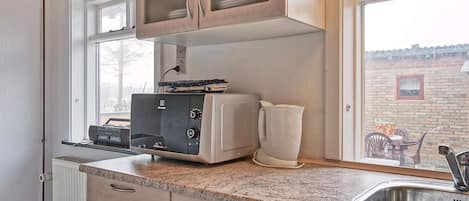 A kitchen with wooden cabinets, a built-in microwave, a kettle, and a window with a view of a brick wall and outdoor furniture.