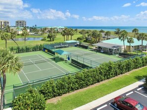 A tennis court with a covered seating area, surrounded by greenery and a view of the ocean.